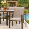 An outdoor patio setting featuring modern bronze-toned Urban stackable sling dining chairs paired with a matching outdoor dining table near a poolside deck, styled with wine glasses, a glass candle holder, and a vase of white flowers.