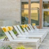 A row of white Urban stackable commercial chaise lounges lined up on a modern concrete patio outside a building, each equipped with bright yellow cylindrical headrest pillows and separated by small white cylindrical side tables.