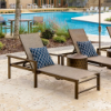 A pair of bronze-toned Urban aluminum chaise lounges with arms arranged on a paved resort pool deck, styled with blue-patterned throw pillows and a round metal side table near a swimming pool.