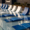 A line of white and blue Marine Grade Polymer slatted chaise lounges partially submerged in clear water on a commercial pool tanning ledge.