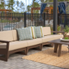 Outdoor patio setup featuring a multi-seat modular commercial sectional with dark frames, light beige cushions, accent pillows, and an outdoor coffee table on a paved stone patio.