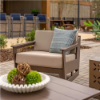 A commercial outdoor cushioned lounge chair with dark brown frame and beige cushions, featuring a decorative throw pillow, styled on a stone patio next to an outdoor table.