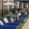 Outdoor patio lounge area set on artificial turf featuring gray wicker chaise lounges with royal blue cushioned tops, matching club chairs, side tables, and a patio umbrella.