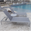 Side view of a round aluminum side table placed between two grey sling chaise lounges on a concrete pool deck next to a swimming pool.