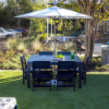 Outdoor lifestyle shot of a dark navy 42x60-inch MGP rectangular dining table set on artificial lawn turf, surrounded by six matching navy chairs and shaded by a large white patio umbrella anchored in the center.