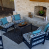 Patio seating arrangement set on a stone hearth in front of an outdoor fireplace, featuring dark-framed MGP deep seating lounge chairs with blue-and-grey geometric patterned cushions, solid teal throw pillows, and a low dark coffee table on a dark outdoor rug.