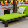 A commercial outdoor pool deck chaise lounge featuring a dark chocolate brown woven wicker frame topped with a vibrant lime green full-length cushion, a pattern accent pillow, and a small dark side table placed alongside.