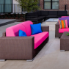 A commercial outdoor two-seat loveseat featuring a dark Java chocolate brown woven resin wicker frame with wide block arms, vibrant hot pink cushions, blue accent pillows, and modern silver feet, set on a stone patio arrangement.