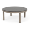 A round patio coffee table featuring a neutral grey slatted MGP tabletop set on a Satin Amber aluminum frame with four straight legs.