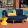 A vibrant yellow diamond-shaped fiberglass side table positioned next to a curved outdoor lounge daybed with navy blue cushions and colorful throw pillows beside a pool deck.